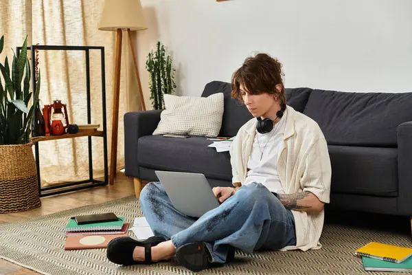 Odaklanmış genç bir öğrenci evde çalışma materyalleriyle çevrili olarak yerde dizüstü bilgisayarıyla meşgul oluyor..