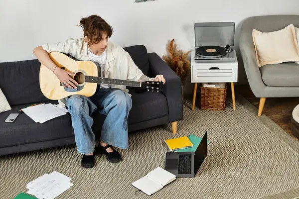 Genç bir öğrenci kanepede gitar çalar. Etrafı kağıtlarla ve rahat bir evde dizüstü bilgisayarla çevrilidir..