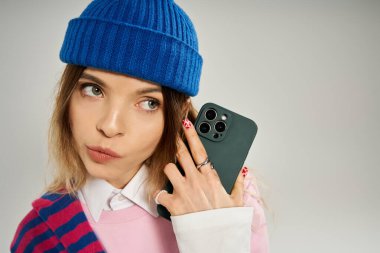 Moda kıyafetleri giymiş genç bir kadın akıllı bir telefon tutuyor ve cazibesiyle poz veriyor..