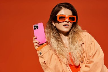 Kendine güvenen genç bir kadın pembe telefonuyla poz veriyor, parlak bir günde göz alıcı bir kıyafet giyiyor..