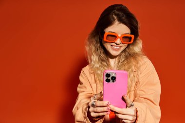 Moda kıyafetli genç bir kadın canlı turuncu bir çevrede selfie çekerken gülümsüyor..