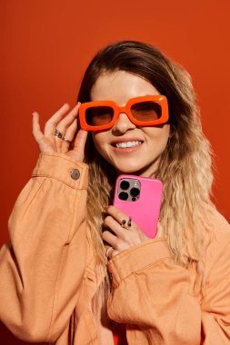 Şık bir genç kadın pembe bir telefon tutarken, canlı giyinmiş bir şekilde gülümsüyor..