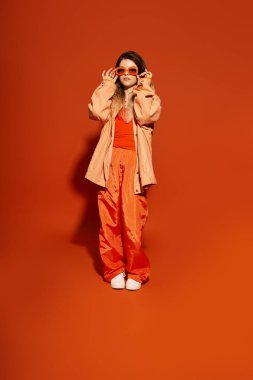 Güzel genç bir kadın canlı turuncu bir arka planda kendine güvenerek duruyor, şık kıyafetini sergiliyor..