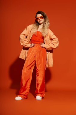 Renkli kıyafetli ve aksesuarlı kendine güvenen genç bir kadın turuncu bir arka plana karşı poz veriyor..