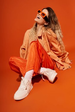 Parlak ve neşeli genç bir kadın şık bir turuncu elbiseyle poz veriyor, neşe ve güven saçıyor..
