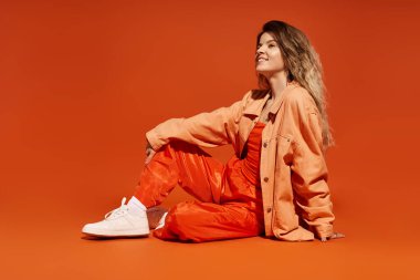 Tarz sahibi genç bir kadın zarif bir şekilde oturur, turuncu bir arkaplana karşı kendine güvenir..