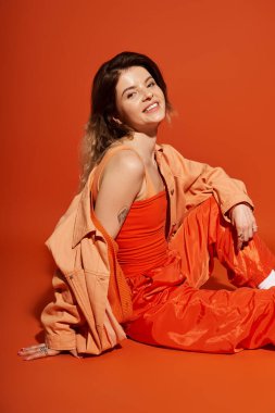 Şık bir kadın şakacı bir şekilde yerde rahatlar, canlı kıyafeti turuncu bir zemine dayanır..