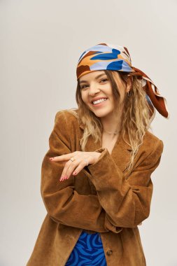 Kendine güvenen genç bir kadın, gri bir arka planda şık bir kıyafet ve canlı bir başörtüsü sergiliyor..