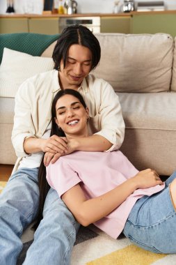 Bir erkek, yumuşak bir kanepede gülümseyen bir kadını nazikçe kucaklar. Evlerinde sıcaklık ve şefkat saçar..