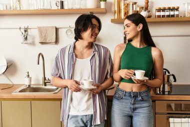 Parlak bir mutfaktaki çift, ellerinde kahve fincanları tutarken gülümseyerek sevgi ve neşe saçıyor..