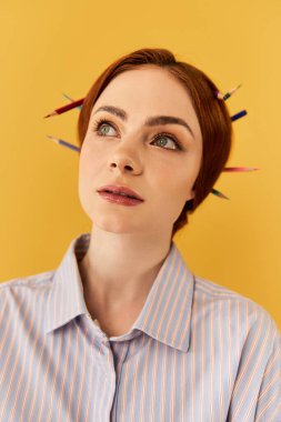 Genç bir kadın, parlak sarı bir duvara karşı, saçlarında renkli kalemlerle yukarıya bakıyor..