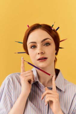 Genç bir kadın parlak sarı bir arka plana karşı saçında kalemlerle poz veriyor..