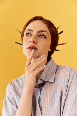 Genç bir kadın, saçlarında renkli kalemler ve parlak sarı bir fon ile düşünceli poz veriyor..