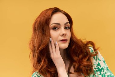 Güzel genç bir kadın dalgalı kızıl saçlarıyla nazikçe yüzüne dokunuyor, özgüvenle gülümsüyor..