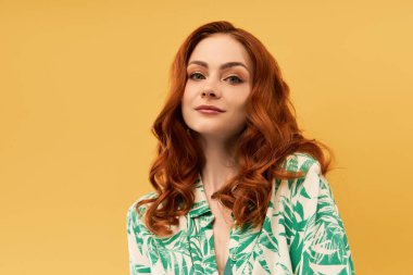 Kendine güvenen kızıl saçlı bir kadın zarif bir şekilde sarı bir arka planda duruyor..