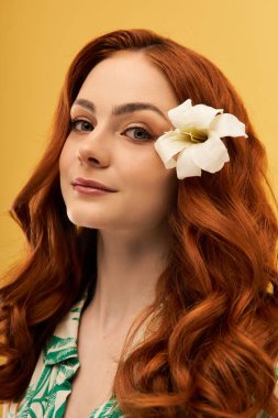 Güzel, genç bir kadın kendine güvenerek duruyor. Kızıl saçları ve kulağında beyaz bir çiçekle..