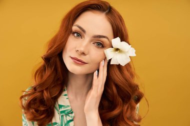 Uçan kızıl saçlı ve beyaz çiçekli genç bir kadın gülümserken yüzüne hafifçe dokunur..