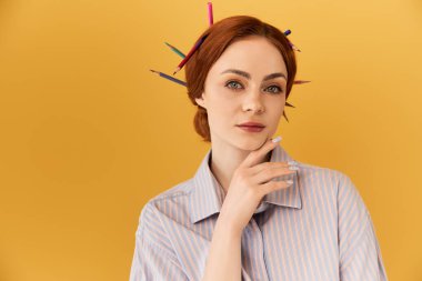 Parlak giyimli kadın, sarı bir duvarın yanında saçlarında renkli kalemlerle sanatsal yetenekler sergiliyor..