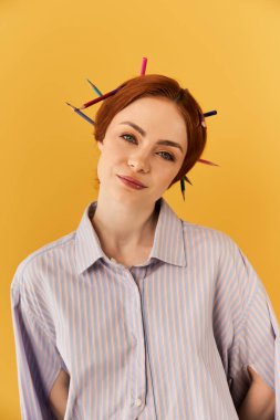 Genç bir kadın saçlarında renkli kalemlerle sarı bir fona karşı neşe içinde poz veriyor..