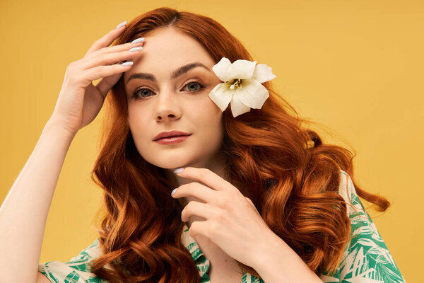Красивая молодая женщина с вьющимися рыжими волосами нежно прикасается к лицу, демонстрируя цветок..