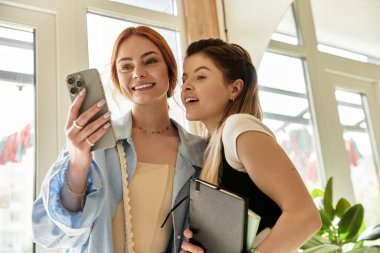 İki şık genç kadın bir kafede keyifli bir anı paylaşıyor, gülümsüyor ve telefona bakıyor..