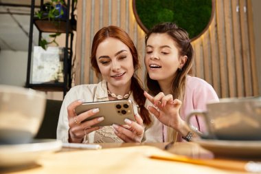 İki genç kadın moda bir kafede telefonlarını karıştırırken keyifli bir anı paylaşıyorlar..
