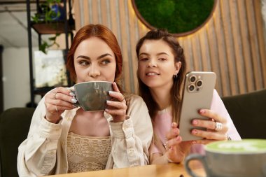 Birbirine eşlik eden iki arkadaş kahvelerini yudumlar ve şık bir kafede gülücüklerini paylaşırlar..