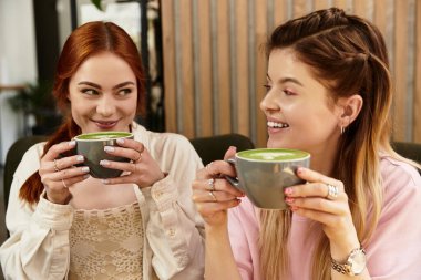 İki genç kadın, modern bir kafe ortamında gülüşlerini ve yeşil içkilerini paylaşıyor, arkadaşlığı kucaklıyor..