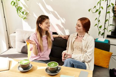 İki genç kadın, sıcak bir kafede yeşil içkileri yudumlarken kahkaha ve hikayelerini paylaşıyor..