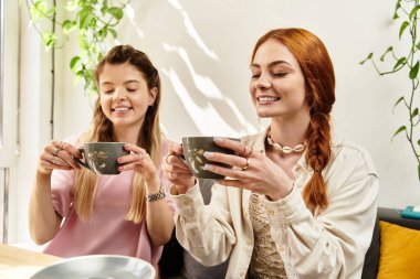 İki genç kadın, yeşilliklerle dolu canlı bir kafede sıcak bir içki eşliğinde keyifli bir anı paylaşıyor..