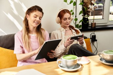 İki genç kadın modern bir kafede rahat bir anı paylaşıyor, cihazlarıyla meşgul..