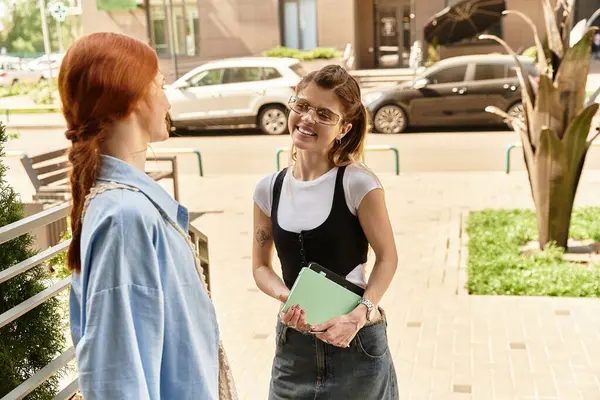 İki şık genç kadın dışarıda gülümseyerek ve bağlı bir şekilde durarak neşeli bir sohbetin tadını çıkarıyorlar..