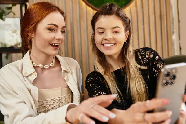 İki arkadaş gülümsüyor ve modaya uygun bir kafede akıllı telefon kullanarak bir anı paylaşıyorlar..