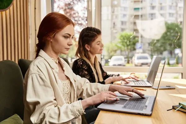 İki genç kadın laptoplarına odaklanırken aynı zamanda canlı bir kafe atmosferinin tadını çıkarıyorlar..