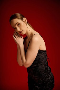 Siyah elbiseli genç bir kadın zarif bir şekilde kırmızı bir arkaplanda poz veriyor..