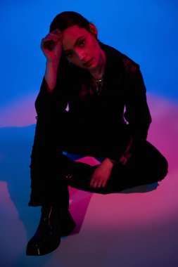 Güzel genç bir kadın zarif bir şekilde pembe ışıkla aydınlatılmış mavi bir arkaplanda oturuyor..