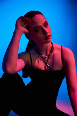 Şık bir stüdyoda, genç bir kadın pembe ışıklandırmalı ve mavi arka planlı modaya uygun kıyafetlerini modelliyor..