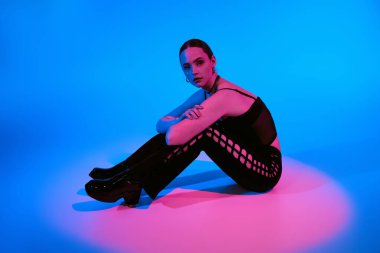 Güzel genç bir kadın, çarpıcı bir mavi ve pembe arkaplan karşısında şık bir kıyafet sergiliyor..