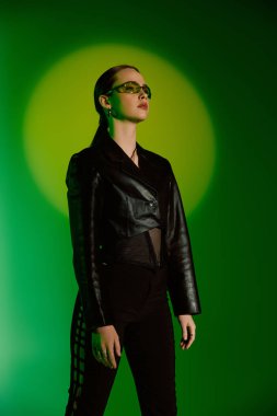 Şık, genç bir kadın kendine güvenerek duruyor, canlı yeşil bir ortamda elbisesini sergiliyor..