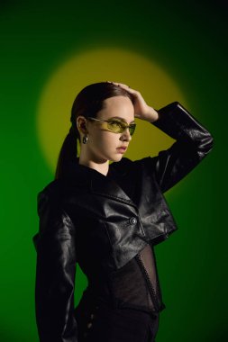 Tarz sahibi genç bir kadın yeşil bir arka planda parlak siyah bir elbiseyle kendine güvenini sergiliyor..