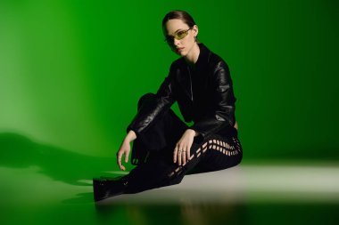 Şık siyah elbiseli genç bir kadın zarif bir şekilde yeşil arka planda oturuyor..