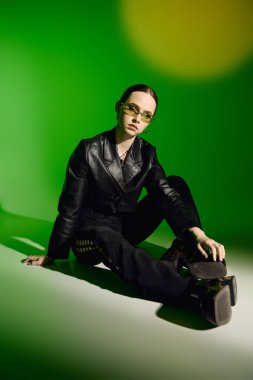 Tarz sahibi genç bir kadın canlı yeşil bir zemine karşı siyah bir elbise içinde poz veriyor..