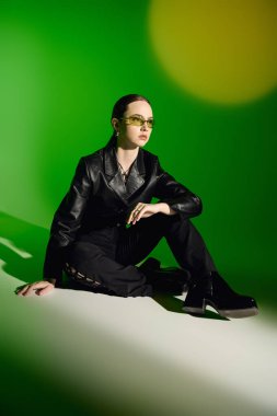 Şık, siyah bir elbise giymiş güzel bir genç kadın parlak yeşil bir arka planda zarif bir şekilde oturuyor..