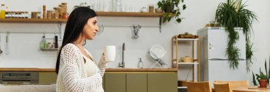 Genç hamile bir kadın sıcak bir içecek yudumlarken güneşli bir mutfakta dinleniyor..