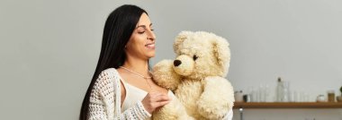 Genç bir kadın neşeli bir şekilde oyuncak ayıyı kucağında tutuyor, rahat bir ortamda hamileliğini kucaklıyor..