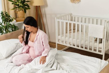 Pembe pijamalı bir anne, yumuşak battaniyeler ve bebek odasıyla çevrili yatakta düşünceli bir şekilde oturur..