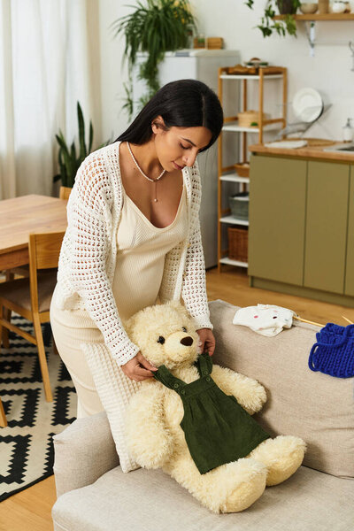 Молодая беременная женщина одевает плюшевого медведя в уютную гостиную для своего новорожденного.