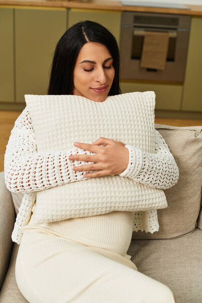 Молодая женщина, ожидающая ребенка, обнимает мягкую подушку, наслаждаясь спокойным моментом в помещении.