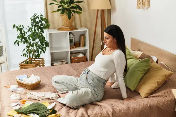 Güzel genç bir kadın yatağında oturmuş sıcak bir atmosferde bebek kıyafetleri ve aksesuarlarını düzenliyor..