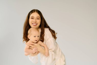 Gülümseyen anne meraklı bebeğini kucaklar, parlak bir ortamda sevgi ve neşe anı yaratır..